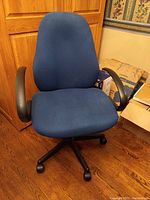 Front angled view of a blue fabric upholstered office chair featuring black armrests and a five-star black base with castors on a hardwood floor near a wooden cabinet.