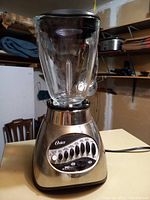 Photo of Oster blender showing stainless steel base with speed control buttons and clear glass blending jar with black lid, on cream countertop in storage room.