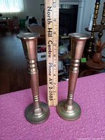Two tarnished brass candlesticks standing on a surface with a ruler next to them showing they are 8 inches tall