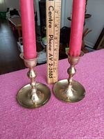 Pair of brass candlesticks with red candles standing on a purple surface with a ruler showing height around 4 inches each.