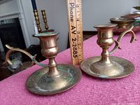 Photo showing two brass candlesticks with handles, one closer to ruler showing about 3.5 inches height, placed on magenta fabric surface. Tarnish and green patina visible.