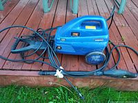 Blue Simoniz pressure washer unit with power cord and integrated hose and spray gun on wooden deck and grass ground