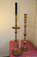 Two tall brass candlesticks on a pink surface with one holding a blue candle, showing segmented construction and a removable tray.