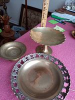 Three brass plates and bowls including a large round plate with cutout edge, medium raised bowl with etched Japanese garden design, and a smaller raised bowl on a table with measuring ruler