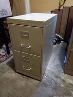 Image of a beige metal two-drawer filing cabinet with metal handles and label holders on each drawer, showing slight wear and tear.