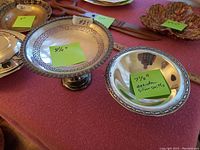 Two silverplate bowls displayed on table with colored price tags. One has intricate cut-out decorative raised edge and pedestal base, size approx. 9.5" diameter. The other is round dish with smooth rim, marked 'Oneida Silversmiths', size approx. 7.5" diameter.