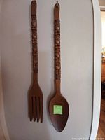 Front view of the large carved wooden tiki fork and spoon hanging on a wall, showing the full length and carved handles.