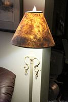 Close-up view of the ornate white metal upper portion of the floor lamp with two curved arms and clear crystal accents hanging below the light socket. The textured amber/brown paper shade is visible.