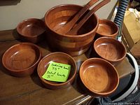 Photo showing the full set of wooden salad bowls, one larger serving bowl with two utensils placed inside, and six smaller salad bowls arranged around it.