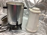 Photo shows 30 cup stainless steel coffee maker with black plastic base and lid along with a white insulated hot/cold beverage carafe.