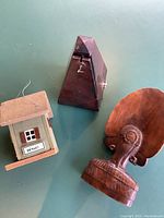 Photo showing three items: a small wooden birdhouse painted to resemble a house, a wooden pyramid-shaped metronome, and a carved wooden bust with feathered headdress.