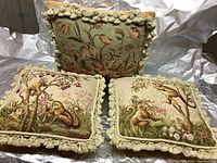 Photo showing three vintage style pillows: two with monkey and floral tapestry designs with fringed edges, and one floral tapestry pillow with pom-pom trim, arranged close together on white surface.