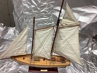 Model sailboat with wooden hull, three fabric sails, and rigging, displayed on a wooden stand with plaque.