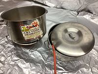 Stainless steel pot with handles open, showing inner marking and an old label about juice extraction. Next to it, the pot lid and a flexible hose for juice extraction.