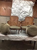 Front view of two antique French provincial chairs with hand cane backs and matching round green cushions on a wrinkled backdrop.