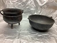 Both the black cast iron scotch bowl/cooking pot with hanging ring and the three-legged cast iron kettle are shown side by side on a silver surface with a silver backdrop.
