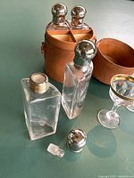 Photo showing four rectangular glass liquor bottles with silver caps, glass stoppers, and one detached stopper alongside a partially open brown leather cylindrical carrying case with compartments.