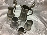 Photo showing five pewter drinking vessels including two large tankards with hinged lids, a medium tankard with an emblem, a plain tankard, and a small two-handled tankard.