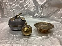 Front view of three decorative brass and pewter table pieces: pumpkin-shaped container, apple-shaped box, and round brass plate.