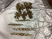Photo showing 4 brass ornate drawer handles with intricate leaf-like designs and 4 smaller brass hardware parts with curvy shapes.