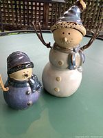 Front view of two ceramic snowmen on green round table outdoors, larger snowman is white and taller while smaller snowman is blue and shorter