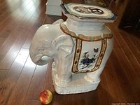 Large white ceramic elephant garden stool with detailed hand-painted decorations, photographed from a side angle showing the design and size compared to an apple on the floor.