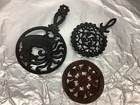Photo of three trivets on a light surface, two cast iron and one wooden, showing detailed designs and round shapes.