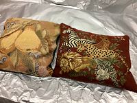 Pair of needlepoint pillows on a silvery fabric background, one with fruit design and the other with exotic animal design