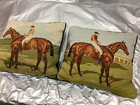 Pair of needlepoint pillows showing English jockeys on race horses, placed side by side on white background