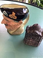 Royal Doulton Monty container or vase depicting a man's head with a black beret, displayed alongside an antique cast iron bank shaped like a house on a green surface.