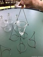 Photo of six clear glass candle holders and their metal wire frames on a table. One holder is suspended from the wire frame.