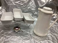 Photo shows three white ceramic rectangular dishes on a metal stand with a small burner below, alongside a tall white ceramic beverage carafe with a lid and handle.