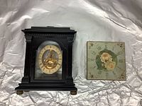 Front view of both decorative clocks: the larger black wooden mantel clock with visible gold pendulum on the left, and the smaller square floral wall clock on the right.