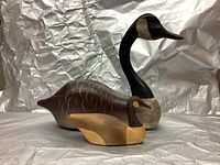 Front angle view of two wood carved decoys: a large goose and a smaller duck, set against a silver crumpled background.