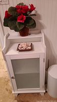 Front view of white wooden bath cabinet with frosted glass door, including faux red flower plant and decorative dish on top.