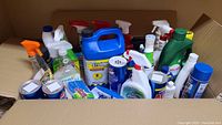 Box filled with various household cleaning supplies, mainly bottles with sprays and liquid cleaners, viewed from above.