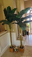Two tall faux green leafy plants in wooden square pots placed indoors next to a stair railing and pillar.
