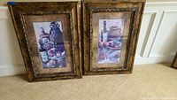 Two framed artwork prints resting against a wall on carpet. Both have rustic ornate brown/gold frames with still life scenes inside.