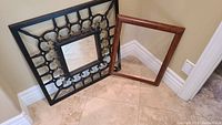 One photo showing both the framed mirror with an ornate metal lattice design and the empty wooden frame with clear glass leaning against a wall on tiled floor.