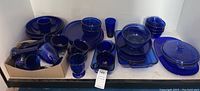 Overview of multiple blue glass kitchenware items including plates, bowls, glasses, and serving dishes arranged on a white shelf space.