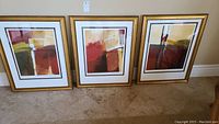 Three gold-framed abstract pictures leaning against a wall on a tile floor.