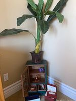Artificial plant atop small dark wooden cabinet containing shelves with a small perfume bottle, artificial purple flower, and several jewelry boxes and wallet on the floor next to it.