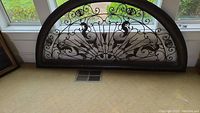 Photo showing arched black metal wall hanging with intricate symmetrical scrollwork, decorative motifs including floral and fan shapes, placed on beige carpet in front of windows.