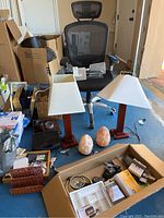Office chair with black mesh back and headrest, two wooden desk lamps with white square shade, two Himalayan salt lamps, and box of various office supplies and tools on blue floor.