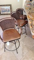 Three brown leather and woven leather swivel bar stools with metal bases shown near a granite counter
