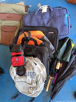 Overview of travel bags including blue laptop bag, beige San Diego backpack, black suitcase, red water bottle holder, umbrellas
