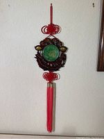 Full view of the Chinese faux jade wall hanging showing the green circular centerpiece, carved dark brown frame with gold characters, red decorative knots, and long tassel.