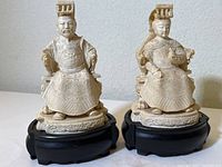 Pair of seated Chinese imperial figures, king and queen, in ivory-toned resin composite on black carved wood bases, showing front view and detailing.