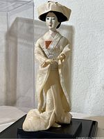 Full view of the 11-inch Geisha figurine in ivory kimono on a black stand inside a clear plastic box.