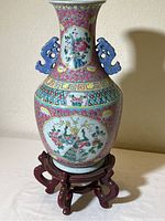 Front view of the tall porcelain vase with floral and bird motifs and blue handles on the neck, shown on the wooden stand.